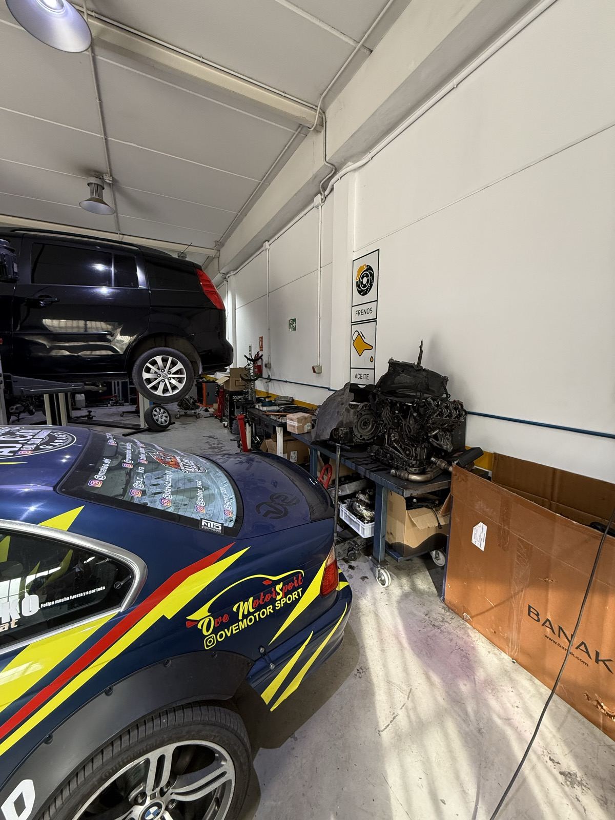 Coche de competición con vinilo azul, amarillo y rojo dentro del taller; al fondo, otro coche en el elevador